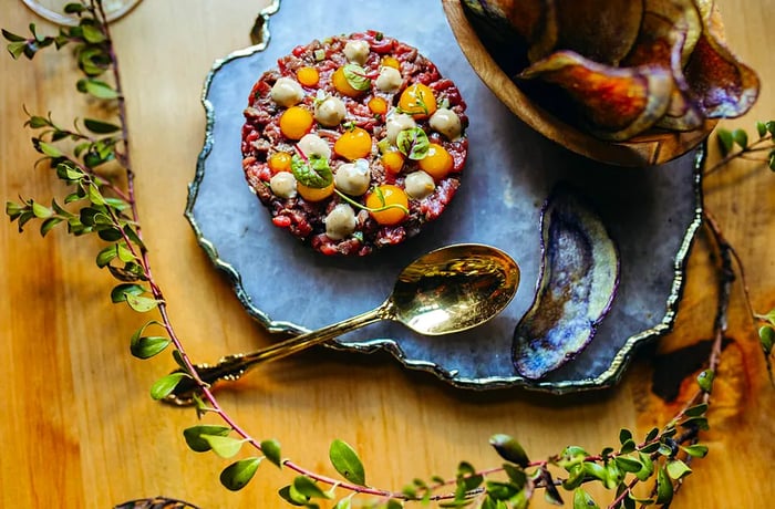 Steak tartare garnished with rich white and yellow sauces, accompanied by dark root vegetable chips.