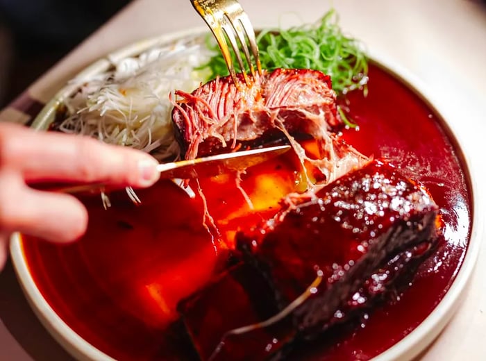 A diner cuts into a beef rib, revealing its juicy interior, served in a rich red sauce alongside rice.