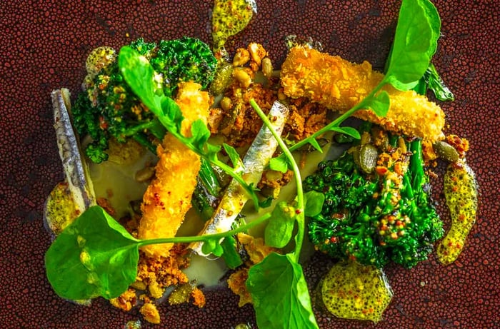 From above, a dish featuring broccoli, sardines, and crispy toppings against a textured backdrop