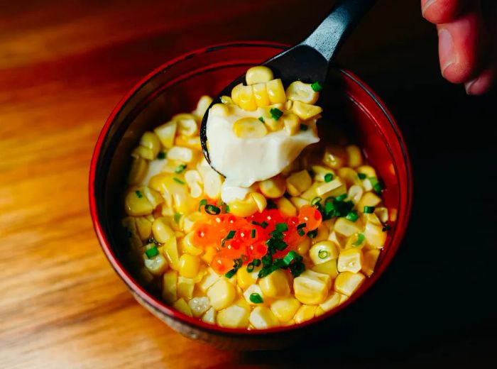 A hand elegantly drizzles cream over a dish filled with corn and roe.