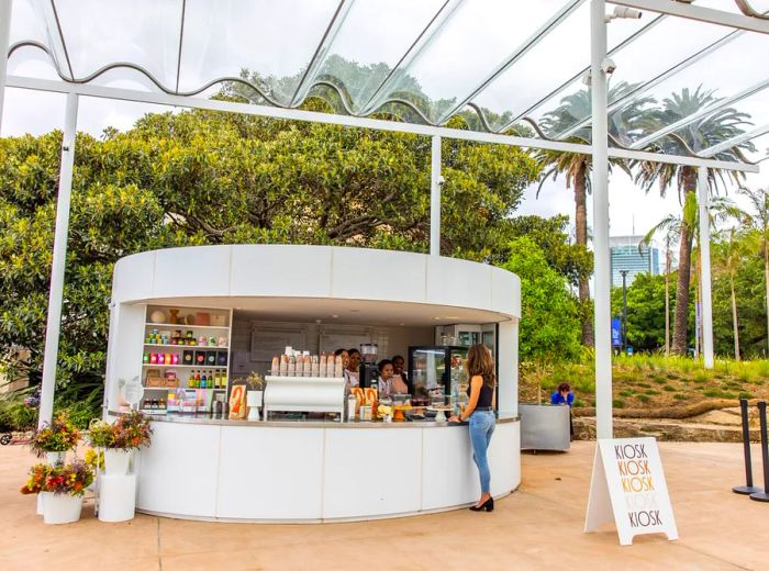 A patron waits at an outdoor kiosk sheltered by a glass awning and greenery.