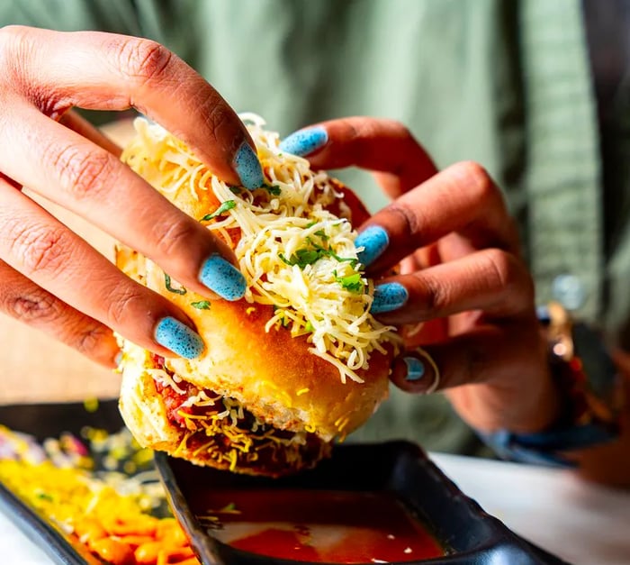 A diner with vivid blue nails lifts a sandwich brimming with onions and cheese, poised above a selection of chutneys for dipping.