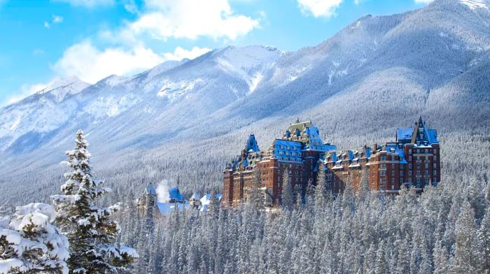 An ornate 19th-century hotel emerges from snow-dusted evergreens, framed by towering mountains in the background.