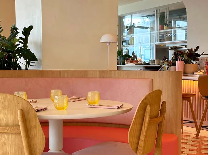 The restaurant's interior features soft hues of pink, red, and white. A banquette table is set with bright yellow cups and sleek modern chairs.