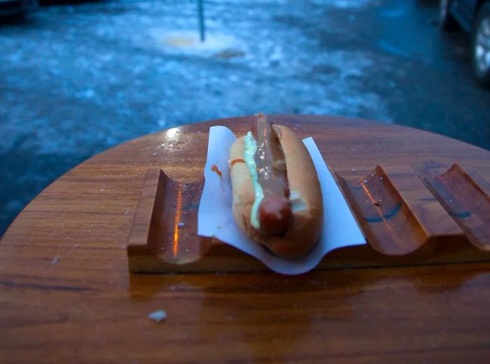 A hot dog nestled in a bun rests in a ridged holder on an outdoor table, with a snowy parking lot and buildings in the backdrop on a cloudy day.