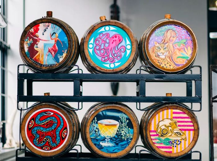 A collection of barrels adorned with various illustrations for aging spirits, displayed on a rack in a stylish industrial setting.