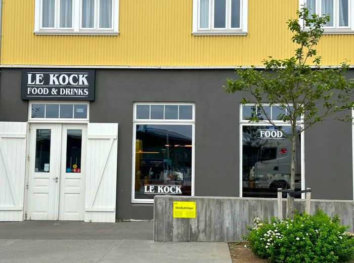 The restaurant's exterior features a neutral palette of gray and yellow, highlighted by large white doors and the establishment's name prominently displayed in various locations.