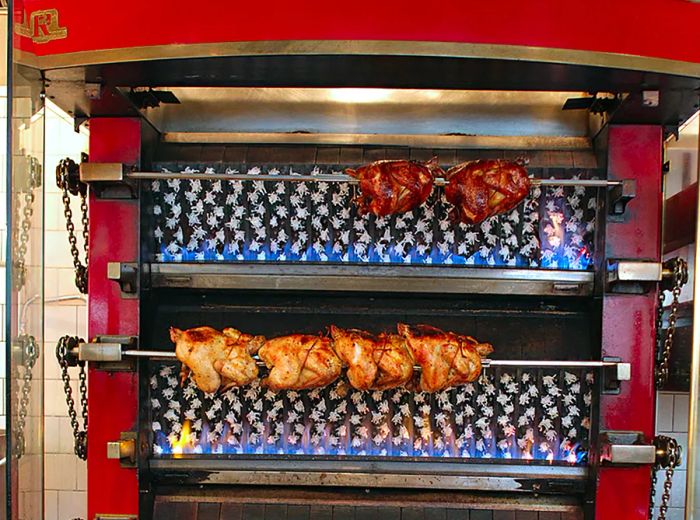 Whole chickens roasting on spits within a large red rotisserie oven.