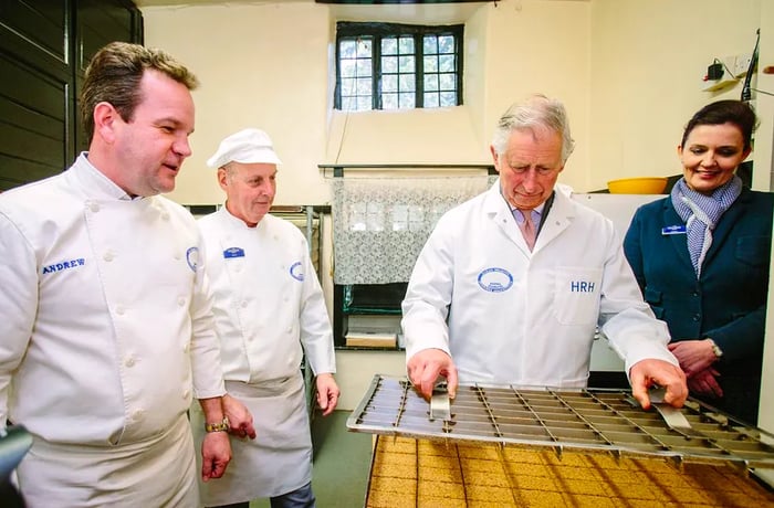 Prince Charles holds a large cutting tool above a tray of gingerbread while bakers observe.