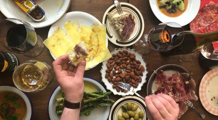 A hand reaches for a piece of bread topped with fish, surrounded by a table adorned with dishes such as asparagus, sliced meats, a tin of fish, thin cheese, olives, and various bottles and glasses of red and white wines.