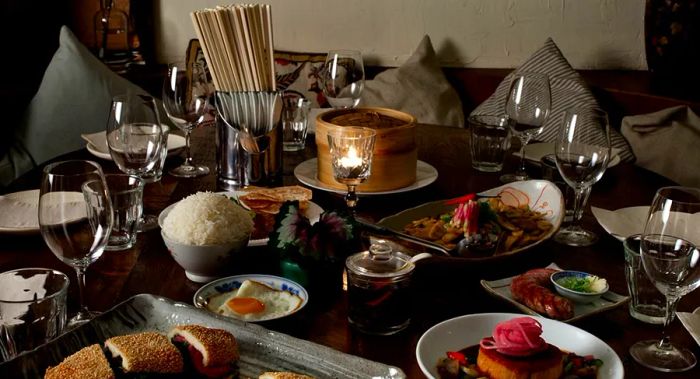 A candlelit table laden with a variety of dishes, a mound of white rice, a fried egg, sliced sandwiches, a bamboo steamer basket, and elegant glassware.