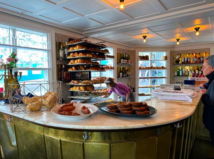 A bakery employee retrieves a pastry from a shelf behind the counter as a customer observes.