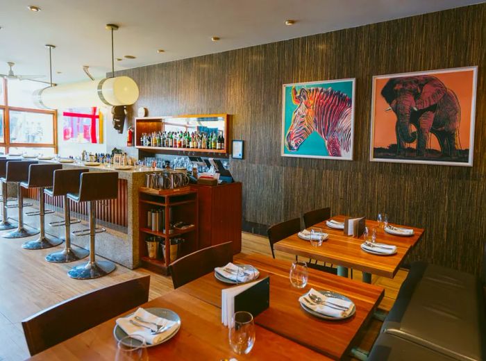 The restaurant features an interior adorned with textured wooden walls, a bar illuminated by a long pendant light, tables elegantly set, and playful illustrations of a zebra and elephant.