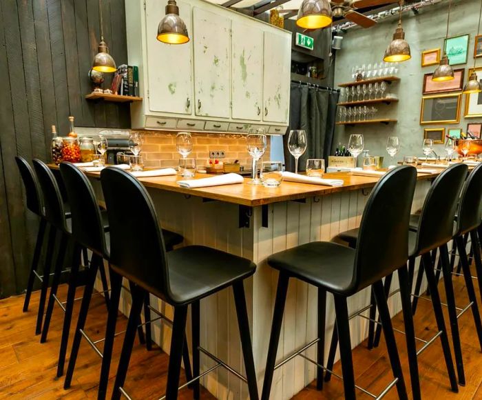 A small counter adorned with table settings, surrounded by bar stools in a rustic industrial atmosphere featuring metal pendant lights, aged kitchen cabinets behind the counter, and an assortment of small pictures on the wall.