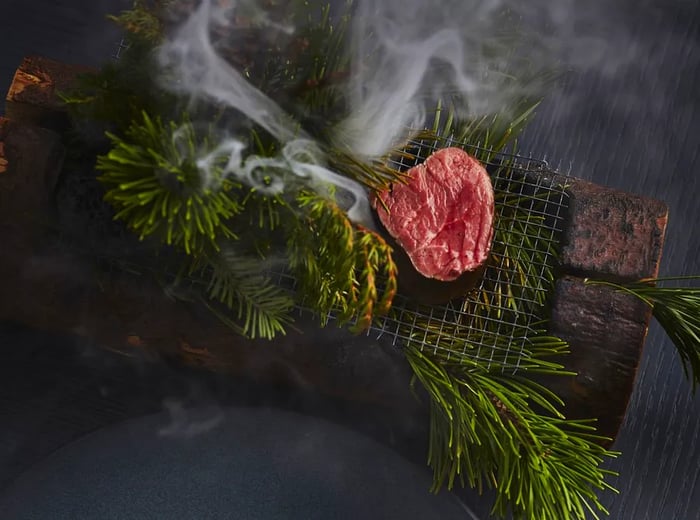 A beef fillet emits wisps of smoke as it cooks on a rustic tabletop grill, surrounded by an assortment of ferns and brush used for smoking.