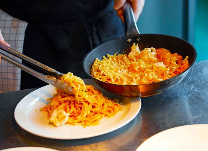 A chef uses tongs to plate a generous portion of pasta with shrimp from a skillet.