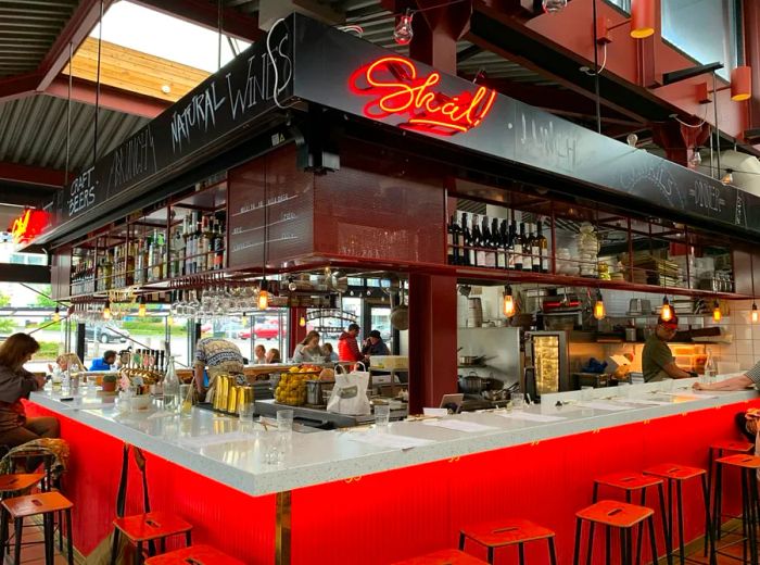 An expansive U-shaped counter encircles a bar and open kitchen, illuminated by a vibrant neon sign within a stylish industrial setting.