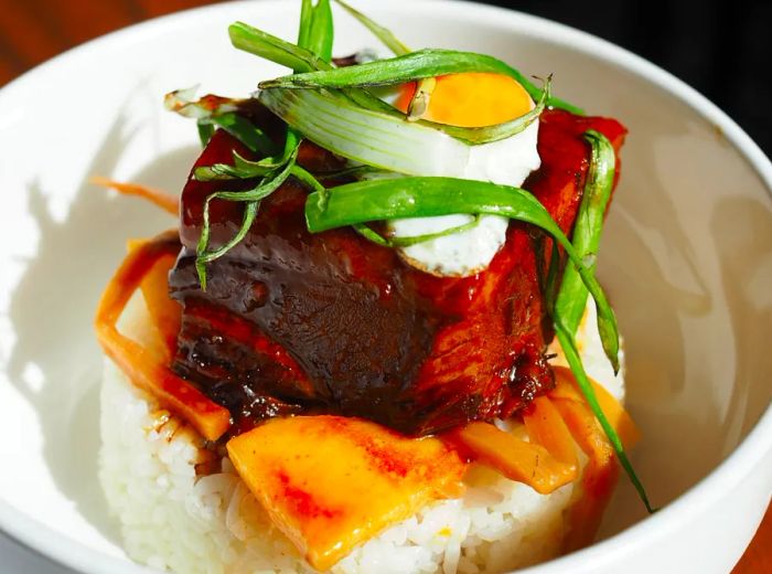 A fried egg perched atop a generous slice of saucy meat, served over rice.