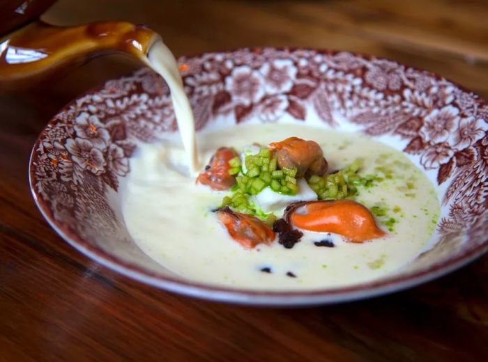 A delicate halibut soup is poured from a kettle into an elegant bowl, already filled with apples, raisins, and mussels.