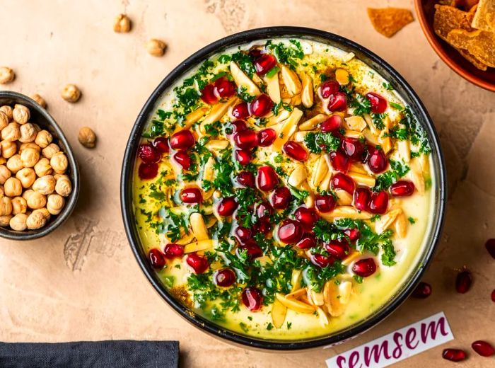 A bowl of fatteh garnished with fresh herbs, pomegranate seeds, and sliced nuts.