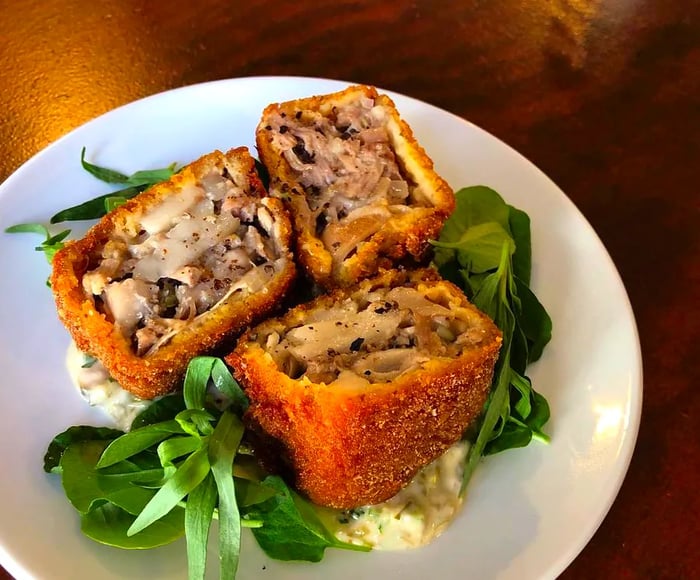 Three pieces of a pastry, split open and filled with meat, cheese, and herbs, resting on a bed of greens with a hearty sauce on a white plate atop a wooden table.