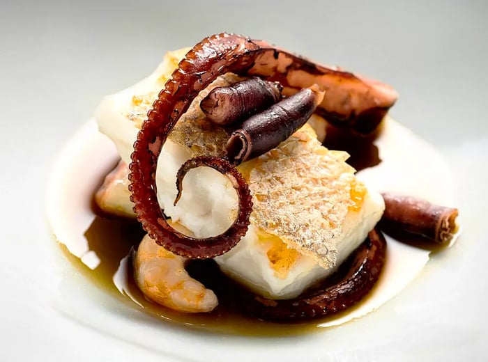 A tentacle of octopus elegantly placed atop a bed of other ingredients in a rich dark sauce, set against a neutral backdrop