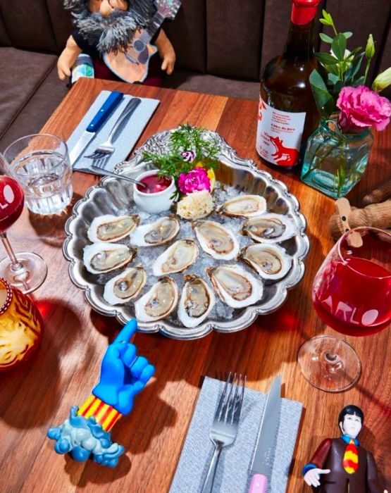 A silver platter holding oysters, featuring a handle resembling a blue genie’s hand, accompanied by glasses of wine.