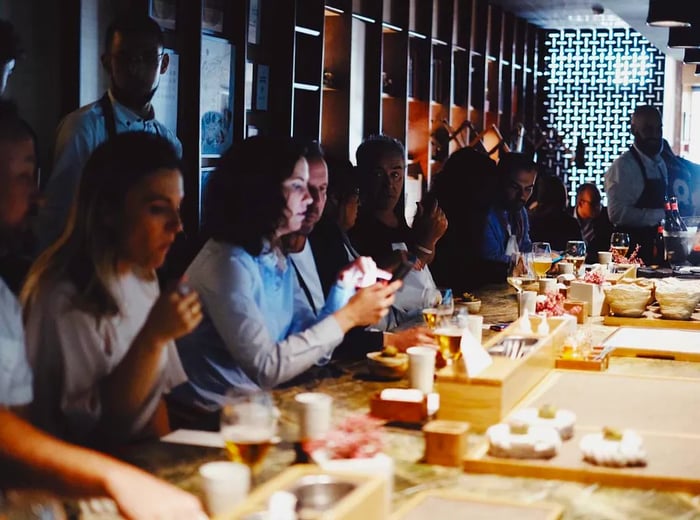 Diners are seated at a glowing chef’s counter, where dishes, slightly blurred, are arranged in various stages of preparation against the backdrop of a dimly lit dining room, with servers visible in the distance.