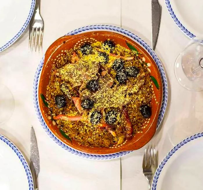 Aerial view of a round plate filled with meat, olives, and vegetables resting on a bed of rice, topped with a sprinkle of cheese, positioned on a white tablecloth amidst four empty place settings.