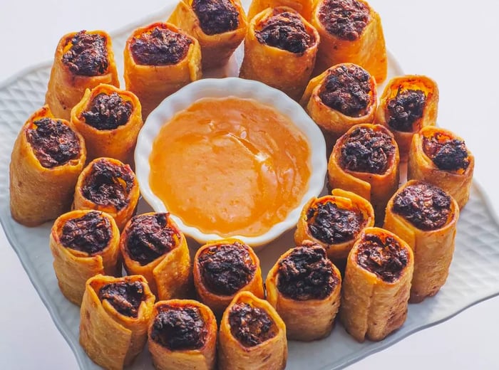 A platter featuring mini egg rolls surrounding a bowl of deep orange dipping sauce.