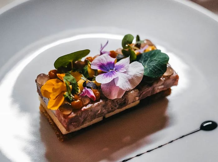 A thick rectangular slice of terrine, adorned with chopped nuts and edible flowers, rests on a white plate beside a delicate streak of sauce.