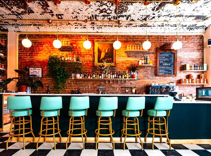 A vibrant bar area featuring checkerboard tile floors, bright blue stools, an exposed brick wall, and stylish orb pendant lights.