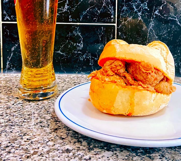 A small sandwich on a plate next to a glass of beer, set on a marble counter against a marble-tiled wall.