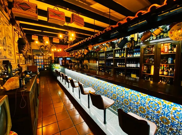 A bar counter adorned with decorative illuminated tiles below, accompanied by bar stools; a well-stocked bar with hanging pots overhead; a tiled floor; walls embellished with framed artwork; and baskets suspended from the exposed wooden rafters.