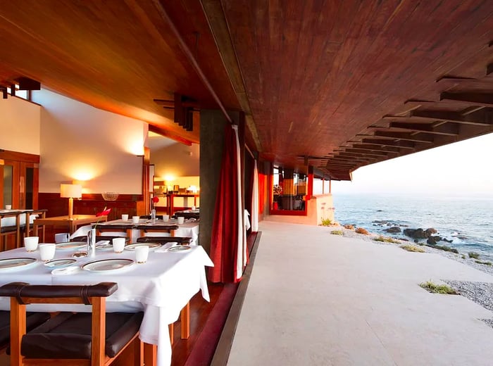 A sophisticated dining area nestled within a striking, curvilinear wooden facade, perched right by the sea with waves crashing on nearby rocks.