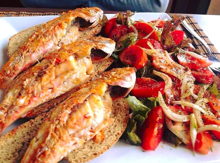 Three whole fried fish are laid on slices of bread on one side of a plate, accompanied by a salad of onions and tomatoes.