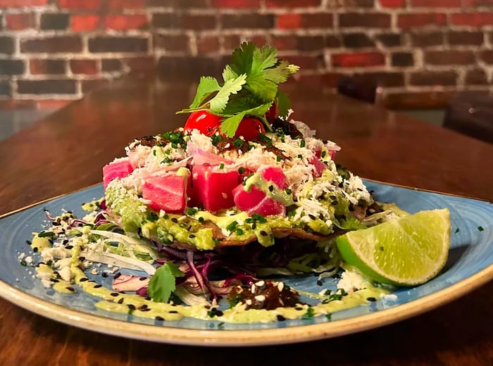 A tostada adorned with large pink fish cubes, topped with cheese and chopped vegetables, accompanied by a lime wedge.