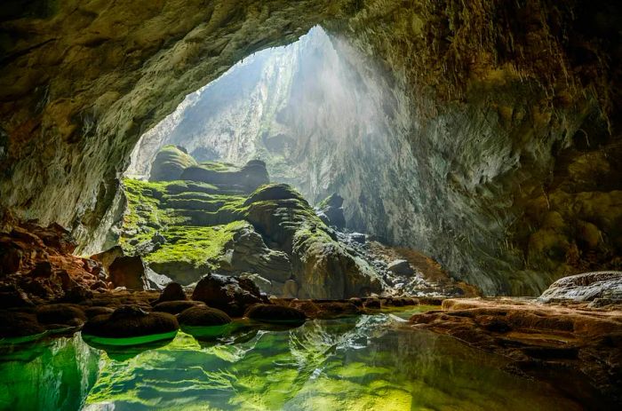 Sunlight pours through an opening in the cave, illuminating a clear green pool of water below.