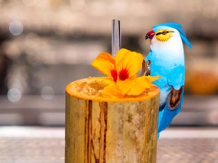 A highball glass resembling wood, featuring a vibrant fake bird perched on the rim alongside a flower garnish and a straw protruding from a drink filled with crushed ice