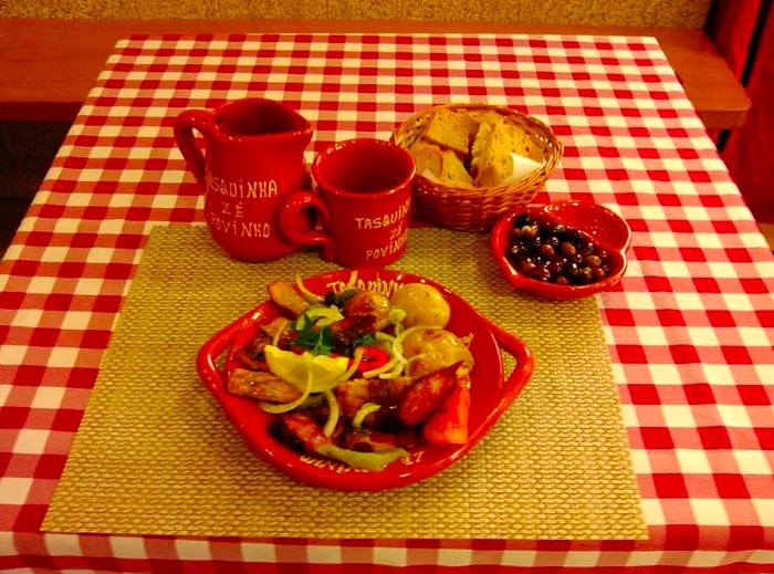 A beautifully arranged plate featuring meat and potatoes, accompanied by a basket of bread and a bowl of olives, set on a checkered tablecloth alongside a mug and pitcher.