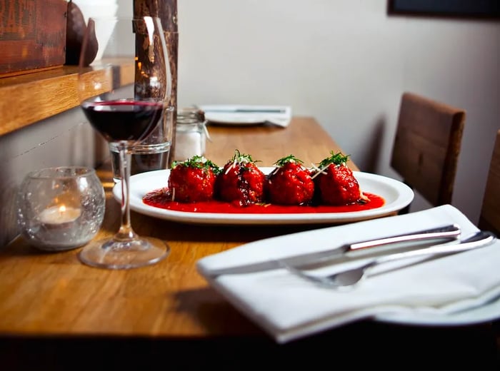 A plate of meatballs drenched in red sauce rests on a bar beside a glass of wine.