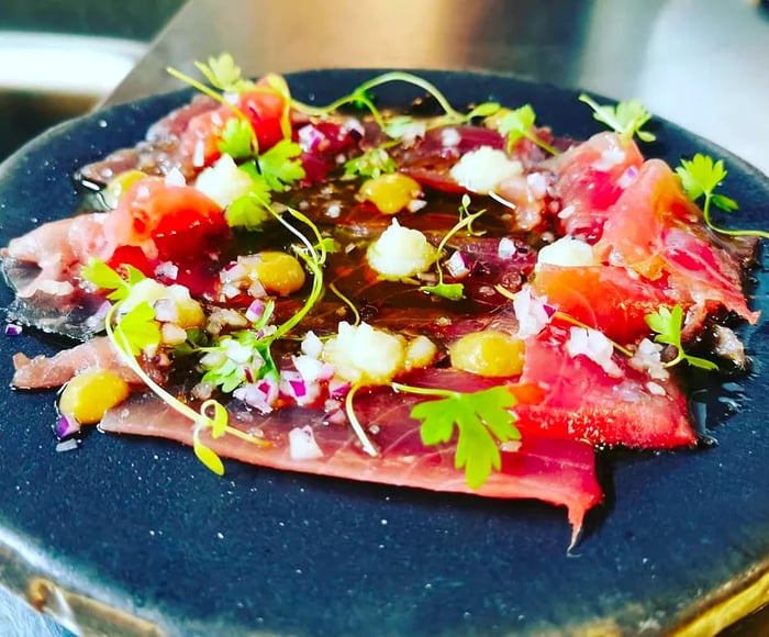 A round, stone-like plate featuring thin slices of fish adorned with a vibrant array of herbs, sauces, and other garnishes.