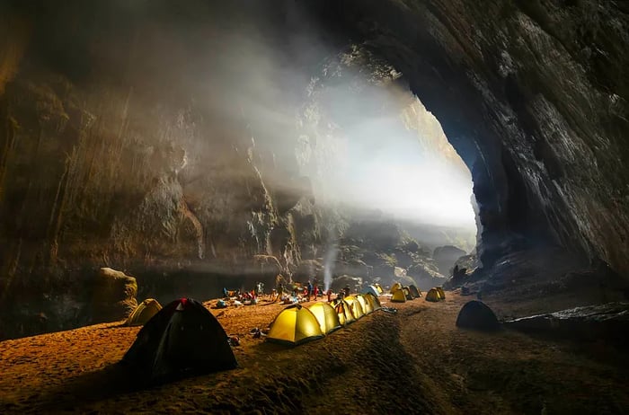 A line of tents is set up on the rocks inside Hang Son Doong cave, with sunlight streaming through an opening.