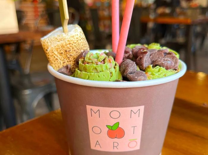 A branded Momotaro cup filled with green tea ice cream, sweet red beans, a torched marshmallow on a skewer, and Pocky sticks.