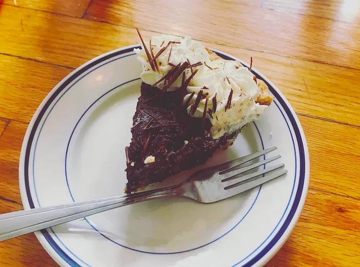 A slice of rich dark chocolate pie adorned with whipped cream and chocolate shavings, accompanied by a fork.