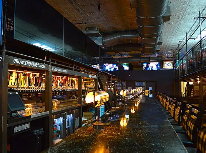 A dimly lit bar featuring a long counter, library-style lamps, rows of bottles on metal shelves, and TVs broadcasting sports overhead.