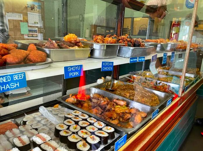 Trays of various dishes displayed in a glass case, labeled with names like Shoyu Pork and Korean Chicken.