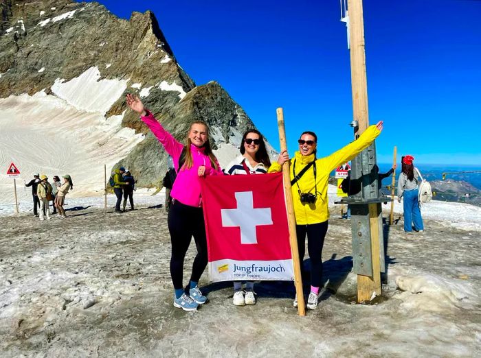 Three women stand atop an alpine peak, proudly holding a Swiss flag.