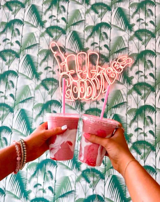 Two hands hold vibrant pink beverages in front of a neon sign that reads 'Good Vibes,' set against tropical palm wallpaper.