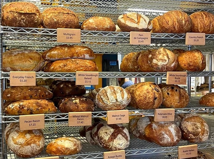 Rows of freshly baked bread loaves neatly arranged on wire shelves, each marked with small labels.